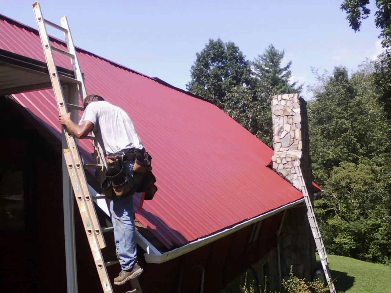 Roofer on a ladder beside a red metal roof and stone chimney — Soffit & Fascia Repair by Roofing Brothers in Center Rutland